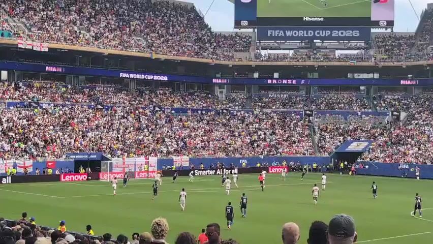 The video shows a wide shot of a crowded soccer stadium during a match. A large scoreboard at the top displays "FIFA WORLD CUP 2026" and a game score, with one team represented by the French flag and the score 0, and the other by the English flag with a score of 3. The field is green with white lines. Soccer players in white and navy blue uniforms are on the field playing. Spectators fill the stands, many wearing white, and a few wearing England flags. The stadium has a roof structure and advertising boards around the field.