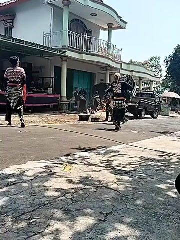 This is a video of a cultural performance outdoors. There are several people involved. In the foreground, a person wearing traditional clothing with a striped shirt, black pants, and a patterned sash walks away from the camera. To the right, another person in similar traditional attire is performing a dance or martial art, with a black mask or headpiece. There are other individuals in the background, some appearing to play musical instruments on a raised platform with red coverings, and others standing near a building. A dark-colored pickup truck is parked to the right. The environment appears to be a street in front of a house with a second story and a balcony. The ground is paved and cracked, with shadows cast by an unseen object. The sky is bright and clear, suggesting a sunny day. The overall scene suggests a local event or celebration.