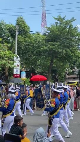 The video shows a marching band in a parade on a sunny day. The band members are wearing blue and gold uniforms with white pants and hats. They are playing brass instruments and marching in formation. There are spectators on the side of the street watching the parade. The background includes trees, buildings, and a tall red and white tower. The camera is filming from a low angle, looking up at the band. In the foreground, the heads and shoulders of several people are visible, indicating they are watching the parade.