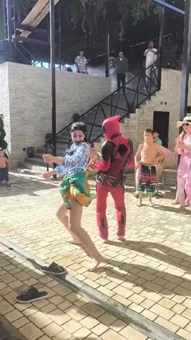 A group of people are dancing outdoors. In the foreground, a woman in a floral top and a green and orange skirt dances. To her right, a person in a Deadpool costume dances. Behind them, several other people watch and dance. The setting appears to be a patio with brick pavers and an overhang.