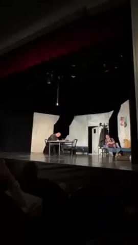 The video captures a scene from a theatrical performance. Two actors are present on a stage with a minimalist set consisting of off-white walls with jagged edges, a table with chairs, and a coat rack near a door. One actor is seated at the table, writing, while the other is sitting on a bench-like structure on the opposite side of the stage, looking towards the first actor. A dimly lit bulb hangs from above. The scene is viewed from the audience, with the backs of heads visible in the foreground. The stage is equipped with several lights. Red curtains are visible at the top of the stage.