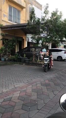 Two women and a child are riding a scooter on a cobblestone street in front of a building. The woman in the front is driving the scooter, and the woman behind her is holding the child. The child is sitting between the two women. The building behind them appears to be a house or a shop, with some plants and trees in front of it. The time of day appears to be late afternoon or early evening, as the lighting is soft. The weather seems pleasant, with no visible rain or strong winds. The scene is likely in a residential or commercial area, given the presence of the building and the paved street. The people appear to be enjoying their ride, with the child looking engaged and the women seeming relaxed. There is no visible text in the media file.