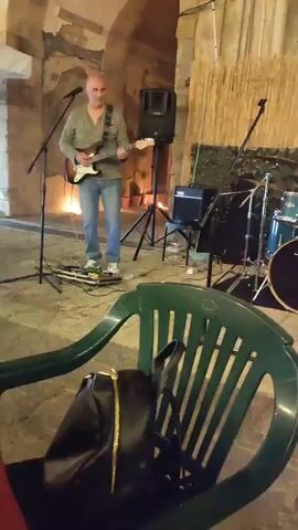 A man plays a guitar and sings into a microphone on a stage. He is wearing a grey long-sleeved shirt and blue jeans. A woman with dark hair stands to his right, singing into a microphone while wearing a black dress and white boots. Behind them is a drum set and a stack of bamboo. In the foreground, a green plastic chair is visible.