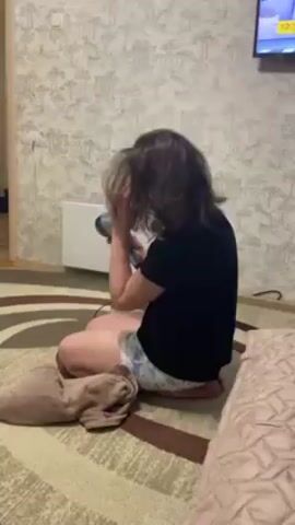 Here's a plain text description of the video:

A woman is sitting cross-legged on a patterned rug, drying her hair with a hairdryer. She is wearing a black t-shirt and patterned shorts. Beside her, a beige towel is folded on the rug. Behind her, a white radiator is mounted on a wall with patterned wallpaper. A television is visible in the background, displaying a program with a time code of 19:30. A portion of a bed is visible to the right of the frame. The woman is facing away from the camera and uses her hand to lift sections of her hair while directing the hairdryer at it.
