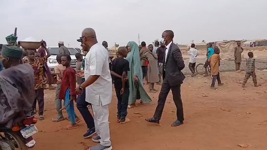 The video shows a group of people, mostly children, gathered in what appears to be a dusty, rural area. Several men, some in formal attire like suits and others in casual clothing, are also present. One person is wearing a face mask. A woman in a pink outfit walks near a black SUV. The background includes simple buildings and a few vehicles, including a white pickup truck and a white van. One child carries a container on his head. The overall scene suggests a community gathering or possibly the distribution of goods in a less developed area.