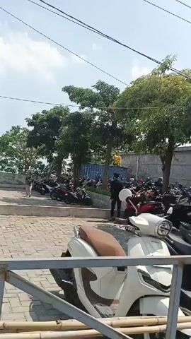 The video shows a parking lot filled with many motorcycles. There are also a few people visible, some walking and some standing. Trees are present in the background, and the sky is clear with some clouds. The ground is paved with interlocking stones, and a metal fence is in the foreground.