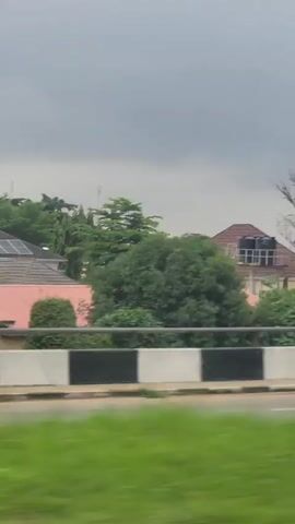 The video shows a view from a moving vehicle. In the foreground, a green grassy area and a white and black striped barrier are visible. A silver Toyota Camry is partially visible on the right side of the frame, with a Nigerian license plate that reads "GWA 418CZ". In the middle ground, there are several buildings with red roofs, lush green trees, and a tall communication tower. The sky is overcast with dark clouds. The scene appears to be an urban or suburban area with residential buildings and infrastructure. The video is shot at an angle, suggesting it was recorded from a car window.