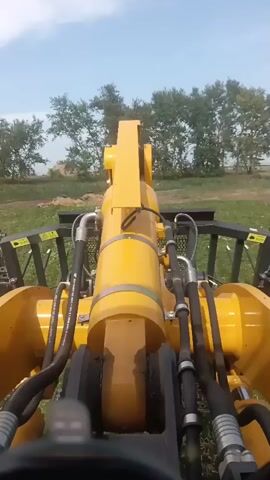 The video shows a first-person perspective of operating heavy machinery. A large yellow hydraulic arm with visible hoses and cylinders dominates the foreground. The arm is attached to a machine with a black metal protective cage visible on either side. Beyond the machinery, a grassy field is seen with scattered trees against a bright blue sky with scattered clouds. The camera appears to be mounted on the machine itself, possibly on the dashboard or operator's seat, providing a forward-facing view of the operation. The overall impression is one of agricultural or construction work being performed in an outdoor, open environment. The scene is static in terms of any human figures, and the primary action observed is the presence and apparent functionality of the heavy equipment.