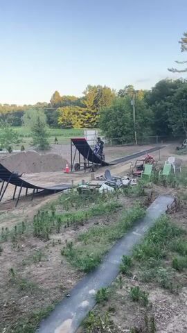 A motocross rider performs a jump from a ramp in a rural setting. The rider is airborne and in mid-air. The background consists of trees and a clear blue sky. Another ramp and a dirt track are visible. The video shows the rider approaching an inflatable landing ramp. In another scene, the rider approaches an inflatable landing bag. The rider approaches and lands on the inflatable.