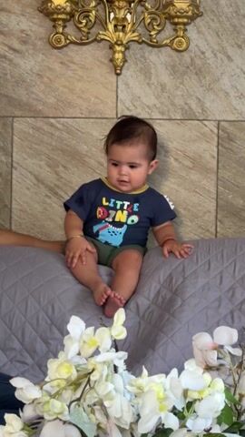 A baby sits on a grey quilted surface in front of a stone-like wall with a gold ornamental piece above.  White flowers are arranged in front of the baby. The baby wears a navy blue shirt with "LITTLE DINO" and a dinosaur graphic, paired with green shorts.  The baby briefly looks away and then back at the camera, laughing and smiling. A hand is visible on the left side of the frame, supporting the baby.