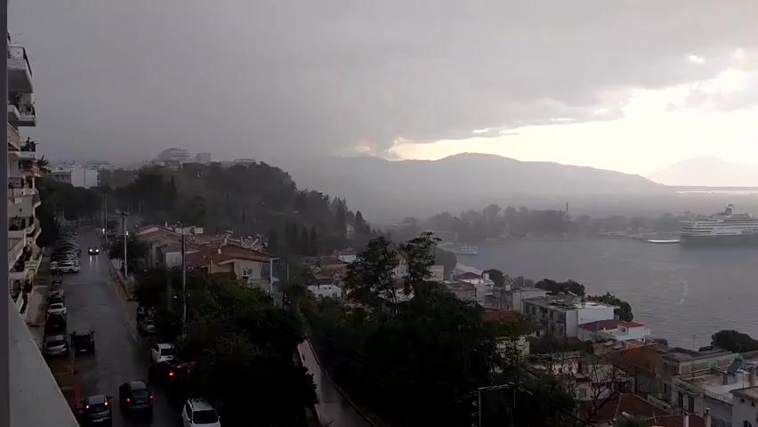 A wide shot shows a coastal town during a heavy rainstorm. The sky is overcast and dark, with streaks of sunlight breaking through the clouds in the distance. Rain is visible falling throughout the scene. In the foreground, a street lined with cars runs alongside a row of buildings with red-tiled roofs. Trees and vegetation are scattered throughout the town. A large body of water, likely a bay or harbor, is visible on the right side of the frame. A large cruise ship is docked in the water. Hills and mountains form the backdrop of the scene, shrouded in mist and rain. The overall atmosphere is somber and dramatic due to the stormy weather.
