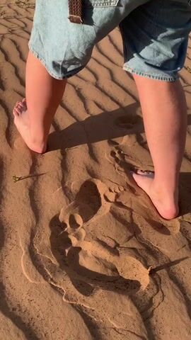 A person wearing denim shorts stands barefoot in a sandy area. The sand has a wavy pattern. The person steps forward, leaving footprints in the sand. The lighting is bright and suggests it is daytime.