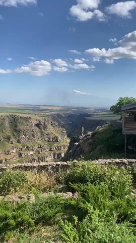 The video pans across a scenic landscape featuring a deep canyon and a snow-capped mountain in the distance. In the foreground, lush green bushes and a stone wall line the edge of the viewpoint. A wooden structure, possibly a cafe or viewing platform, is partially visible on the right. The canyon walls are steep and rugged, with visible layers of rock formations. In the far distance, Mount Ararat stands majestically, its peak covered in snow. A plume of smoke rises from the foothills of the mountain, suggesting potential activity below. The sky is a clear blue with scattered white clouds, indicating a pleasant day. The camera movement is slow and steady, allowing the viewer to take in the expansive vista.