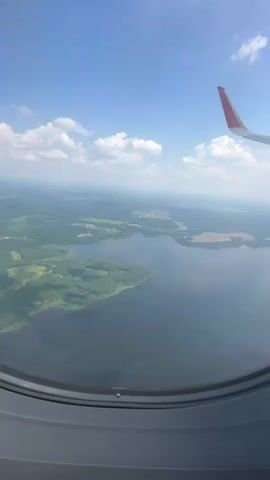 This video shows an aerial view from an airplane window, looking out at a vast landscape. The primary subjects are the natural scenery below and the airplane's wing visible in the upper right corner. The scene is dominated by a large, dark body of water, likely a lake, with a forested shoreline. Beyond the water, the land is a patchwork of green fields and wooded areas, with hints of scattered settlements or buildings in the distance. The sky is a bright, clear blue, dotted with fluffy white cumulus clouds, suggesting a pleasant daytime with fair weather.

The activity is the flight itself, as evidenced by the view from the airplane and the presence of the wing. The time of day appears to be midday, given the bright sunlight and clear sky. The location cues, such as the type of landscape (forests, lake, agricultural fields) and the architectural style of distant buildings, suggest a rural or semi-rural setting. There is no visible text in the video. The overall emotion conveyed is one of travel and expansive beauty, with a sense of peace and tranquility from the elevated perspective. The view offers a sense of scale and the vastness of the natural world.