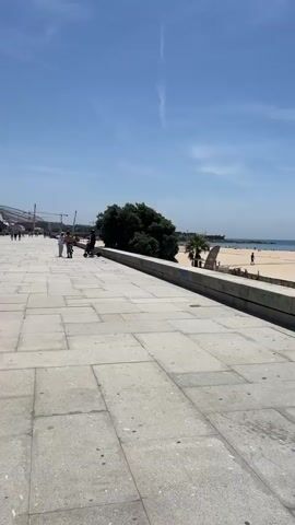This is a vertical video taken from a paved walkway overlooking a beach. The walkway is made of large, light-colored square tiles. A low concrete wall separates the walkway from the beach. On the beach, there are people, umbrellas, and a lifeguard stand. The ocean is visible in the background, with a clear blue sky above.

In the foreground of the video, a person wearing a black baseball cap and a backpack walks away from the camera, their back to the viewer. They appear to be shirtless. Further down the walkway, a group of people are walking towards the camera. One person is on a scooter. To the left of the walkway, there is a large, dark green tree. On the right side of the walkway, near the concrete wall, there is a black trash can.

The video pans slightly to the right, revealing more of the beach and the ocean. A crane or some kind of construction equipment is visible in the distance on the right side of the beach. The sun appears to be shining brightly, casting shadows. The overall impression is a sunny day at a beach resort.