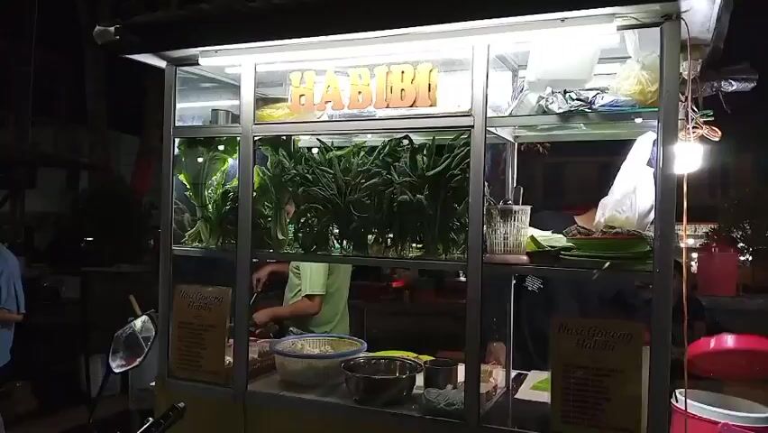 The video shows a food cart at night. The cart has a sign that says "HABIBI". Inside the cart, there are green chili peppers displayed in a glass case. A person wearing a green shirt is cooking behind the counter. There are bowls of ingredients and a sign with a menu in front of the cart. In the foreground, a motorcycle is parked. The background is dark, suggesting it is nighttime. There are also some lights and other structures visible in the background.