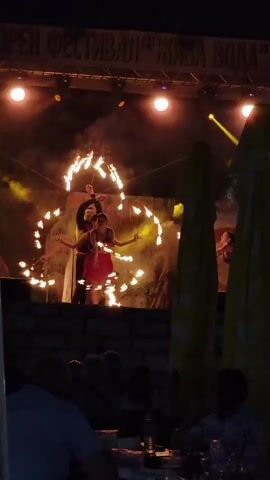 The video shows an outdoor night performance on a stage. The stage has a backdrop and is lit by stage lights. Several performers are on the stage, some of whom are using fire in their act. One performer in a red dress is in the center, twirling flaming props. Around her, other performers in darker clothing are also twirling flaming props, creating a circular effect of fire. The audience is seated in front of the stage, with their backs to the camera, illuminated by the stage lights. The environment appears to be a festival or outdoor event, with umbrellas visible in the background on the right.