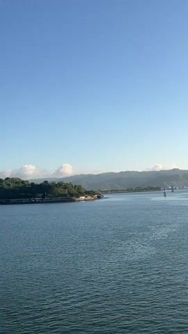 A wide shot captures a calm blue ocean with a distant, tree-covered island on the left. The island features a few buildings nestled among the trees. In the background, rolling hills stretch across the horizon under a clear, bright blue sky with a few scattered white clouds. The water is slightly rippled, reflecting the sunlight. Two faint vertical objects, possibly buoys or small structures, are visible in the water further out.