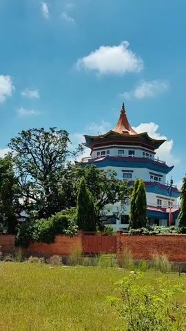 This is a vertical video of a multi-story building with a distinctive pagoda-style roof. The building is white with blue and red trim. It is surrounded by lush green trees and a brick wall. The sky is blue with scattered white clouds. The video pans slightly from left to right, showing more of the building and its surroundings.