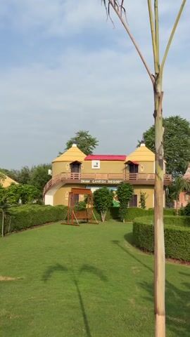 A vertical shot pans across a resort. The resort features several dome-shaped buildings with yellow walls and thatched roofs. The buildings are arranged around a central courtyard with manicured hedges and lush green grass. A wooden swing set is visible in the foreground, and a tall, thin tree stands in the right of the frame. The sky is a clear blue with a few scattered clouds. The overall impression is one of tranquility and relaxation.