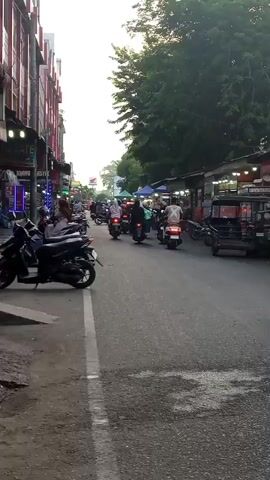 The video shows a street scene with many motorcycles. On the left side of the street, there are buildings with shops. On the right side of the street, there are also shops or stalls with awnings. Many people are riding motorcycles, some with passengers. The traffic appears to be moving slowly. The sky is visible and appears to be a light color, possibly indicating either daytime or late afternoon. There are trees on the right side of the street. The road is paved. There are several parked motorcycles on the left side of the road. One of the shops on the left has neon signs. A small three-wheeled vehicle is visible on the right side of the road. A dark-colored vehicle, possibly an SUV, is entering the frame on the far right.