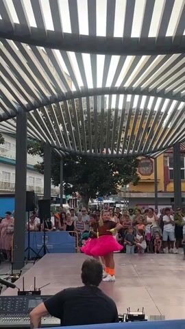 Two young girls wearing bright pink tutus and brown tops are dancing on a stage. A crowd of people watches them from behind a blue barrier. A man in a black shirt is visible in the foreground, operating a soundboard. The stage is set up outdoors, under a pergola with a slatted roof. Buildings and trees are visible in the background. The overall atmosphere appears to be a public performance or event.