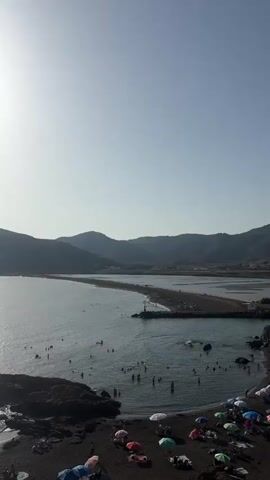 This is a vertical video of a crowded beach with a narrow strip of land extending into the water. The sun is high and bright, casting a glare on the water and illuminating the sky. In the foreground, the beach is dark and appears to be made of small stones or sand. There are several people on the beach, some sitting under colorful umbrellas and others walking. The water is calm and a deep blue, with many people swimming and playing in it. In the middle ground, the strip of land juts out into the sea, and there are more people gathered on it. A small lighthouse or marker stands on the land. In the background, rolling hills or mountains rise up from the coastline. The overall impression is a busy, sunny day at the beach.