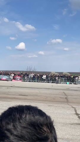 The video captures a drag race between a red car and a blue car on a paved track. A large crowd is gathered behind a metal barrier, watching the event. A man wearing a black hoodie labeled "V.T.A Security" is standing on the track, observing the race from behind. The cars speed off, kicking up dust as they accelerate away from the starting line. The sky is blue with scattered clouds, and a green field stretches in the background beyond the crowd.