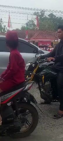 A convoy of motorcycles, some carrying multiple passengers, travels along a dirt road. The motorcycles are diverse in style, with some appearing to be scooters and others more rugged, off-road models. Several individuals are visible on the bikes, including men, women, and at least one child. Many of the passengers are wearing headscarves, and some are dressed in white. Indonesian flags are prominently displayed, attached to poles and some of the vehicles, suggesting a festive or patriotic event. In the background, cars and buildings are partially visible, and the sky is overcast. The road appears to be unpaved, with loose gravel and dirt. The overall impression is of a procession or parade.