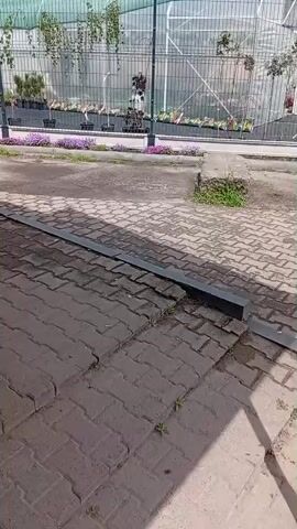 The video shows a paved sidewalk next to a road with a sign in the background advertising a garden center. The sidewalk is made of gray brick pavers and has a black metal border. There is a red metal post in the foreground, partially obscuring the view. The sign in the background is green and white and advertises "Sadovi Dvoryk" (Garden Courtyard) with phone numbers. There are also plants and greenery visible behind a fence in the background. A person is walking on the sidewalk to the right.