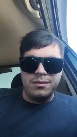 A man with dark hair, a beard, and dark sunglasses is seen from the chest up, seated in what appears to be a car interior during daylight. He wears a dark blue t-shirt and a silver ring on his left ring finger. He looks directly at the camera, raises his left hand to display a peace sign, then lowers his hand.