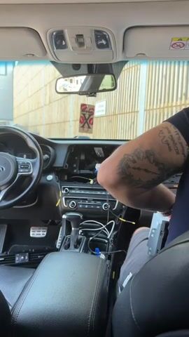 The video shows the interior of a car, specifically the dashboard area. A person with a tattoo on their arm is visible, leaning forward and working on the car's electronics, likely installing or repairing a car stereo. The car's steering wheel, gear shift, and part of the center console are also visible. Wires and electronic components are exposed in the dashboard opening. The background outside the car suggests an outdoor setting, with a wooden-slatted wall visible through the windows. The overall scene depicts a hands-on car maintenance or modification task.