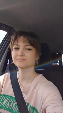 A young woman with brown hair and bangs is seated in the front passenger seat of a car, singing along to music. She is wearing a light pink t-shirt with green lettering and a dark seatbelt is secured across her chest. She has a small stud earring and a hoop earring in her left ear, and a thin gold chain around her neck. Her head is tilted slightly to the right, and her mouth is open as if singing. A faint rainbow-colored light streak is visible on the car door frame next to her. The interior of the car appears to be light-colored, and sunlight is visible through the windows.