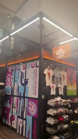 The camera pans to show a retail space called "Local Soul," located within a larger shopping mall. The space features racks of T-shirts, artwork, and faux flowers. A woman with dark hair, wearing a floral skirt and pink top, is browsing the flowers.  Other people are walking by in the mall corridor. The mall features high ceilings and bright, modern lighting.  A sign reading "LEARN" hangs vertically in the distance.  An automated external defibrillator (AED) is mounted on a white brick column.