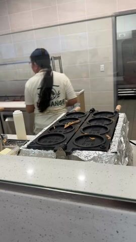 The video shows a woman making pancake-like treats in a commercial kitchen. The woman has long dark hair in a ponytail and wears glasses, a cream-colored t-shirt, jeans, and an apron. The t-shirt has a logo on the back. She works at a counter with various containers and tools. Visible ingredients include chopped cheese, chocolate chips, and a brown powder. A specialized waffle iron type pan sits on the counter, covered in aluminum foil. The pans are black and have circular indents. Steam rises from the pan. The woman walks around the counter and picks up a green bottle. The kitchen has stainless steel appliances and tiled walls. The countertop is a speckled white material.
