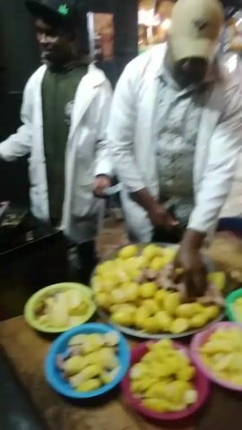 Here's a detailed description of the video:

**Setting:** The video appears to be taken at a small roadside food stall or outdoor restaurant at night. The area is lit by overhead lights and street lights.

**People:** Several people are present. There are two main figures who seem to be preparing food. They are wearing white lab coats, likely as kitchen apparel. Other people are standing around the stall, seemingly as customers.

**Objects:**

*   **Food:** The primary food item visible is boiled potatoes. A large platter is piled high with whole, peeled yellow potatoes. Several smaller, colorful plastic bowls contain cut-up portions of the potatoes. Pieces of meat are included among the whole potatoes.
*   **Cooking Equipment:** There is a multi-burner gas stove with several woks or pans on top. One of the men is using a large spoon to stir the contents.
*   **Counter:** A wooden counter serves as the prep station. It holds the platters and bowls of potatoes.
*   **Other:** A display case, likely for sauces or other condiments, is visible behind the counter. A sign indicating payment services via mobile money is also visible. Several parked cars are visible in the background.

**Actions:**

*   The man in the front is seen cutting up the boiled potatoes on a wooden cutting board.
*   The other man is actively cooking something in the woks on the stove, stirring with a spoon.
*   Customers are approaching the counter, presumably to order food.

**Interactions:** The cooks are focused on preparing and serving food. The customers seem to be interacting with them to place their orders.

**Environment:** The atmosphere appears lively with people and activity, suggesting a popular spot for a quick meal. The lighting indicates it is nighttime. Cars driving past add to the urban environment.