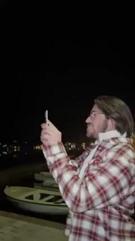 The video shows a man in a red and white plaid shirt and glasses taking a photo with his phone at night. He is standing next to some white rowboats and a short wall. In the background, there are faint lights from what appears to be a town. The sky is dark.