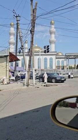 A low-angle shot captures a street scene dominated by a mosque with white minarets and golden domes. Overhead, a dense tangle of power lines crisscrosses the sky. In the foreground, a wooden pole stands prominently, supporting some of the electrical infrastructure. To the left, a small structure with a corrugated metal roof is visible, with bags of goods piled beside it. A few people are present, one walking across the street in front of a dark-colored sedan. The road surface is asphalt, and the sky is clear and blue. The reflection of a side-view mirror can be seen in the bottom right corner of the frame.