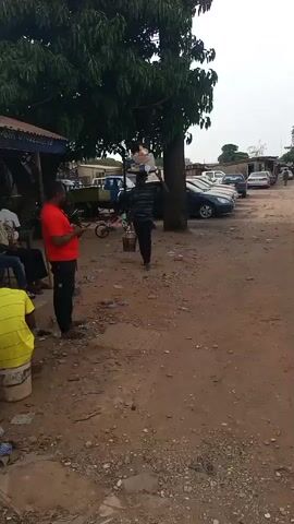 The video shows a man walking on a dirt road. He is carrying items on his head and a container in his left hand. He is walking toward the camera. A man in a red shirt is standing to the left of the road, looking at his phone. There are several cars parked on the other side of the road. Other people are sitting near a building on the left side of the road. There is a tree in the foreground. The setting is outdoors.