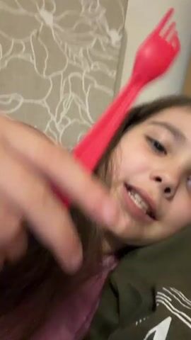 A young girl with long brown hair lies on her side, holding a red plastic fork in her right hand. She brings the fork towards her mouth, then touches her forehead with it. She smiles at the camera. The background shows a wall with a floral-patterned wallpaper.