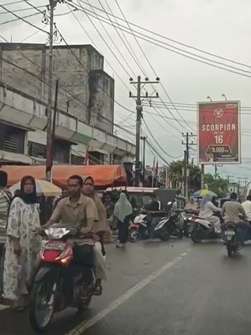 The video shows a street scene with moderate traffic, primarily consisting of motorcycles and a few cars and trucks. The sky is overcast, suggesting cloudy weather. On the right side of the road, there is a billboard advertising "SCORPION 16." The street is lined with buildings and shops, some of which have awnings or temporary stalls. Several people are visible, mostly on motorcycles, with some individuals walking or standing near the shops. The overall impression is that of a busy urban or semi-urban environment during daytime.