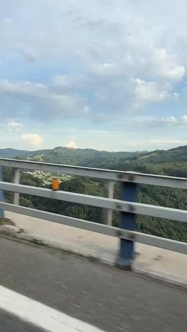 The video shows a view from a moving vehicle on a bridge. The camera is angled to capture the railing of the bridge and the landscape beyond. The railing is made of metal with blue vertical supports and silver horizontal bars. The landscape consists of rolling green hills covered in trees, with some houses and fields visible in the distance. The sky is partly cloudy with a bright blue sky. The white lines of the road are visible in the foreground, indicating the vehicle's movement. There are no people visible in the video. The overall impression is that of a scenic drive through a rural area.