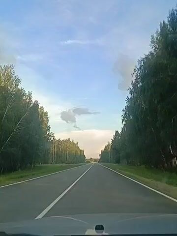 The video shows a forward-facing view from inside a vehicle traveling on a paved road lined with trees on either side. The sky above is blue with scattered clouds. The road appears to be a two-lane highway with a white line marking the center. The vehicle's dashboard is partially visible in the foreground. The time stamp indicates the video begins at 00:00 and ends at 00:04.
