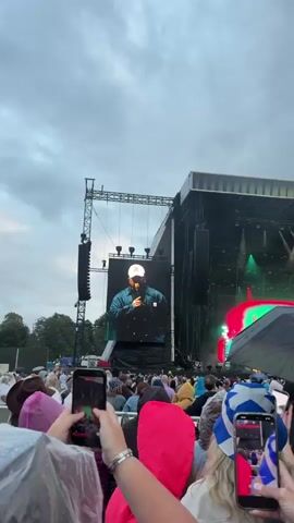The video shows a live outdoor concert. A large screen displays performers. The first performer is a male wearing a white baseball cap and a denim jacket, holding a microphone. The second performer is a female wearing glasses and a black hoodie, also holding a microphone. The screen displays colorful, abstract graphics behind her. A crowd of people, many holding up their phones to record, is visible in the foreground. The sky is overcast.