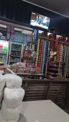 A video displays the interior of a small shop, possibly a convenience store or a kiosk, packed with various goods. The shelves are lined with numerous items, primarily packaged snacks and beverages. In the foreground, there are large white plastic bags. In the background, a television screen shows a news report with three individuals seated at a table. The overall impression is one of a cluttered but well-stocked retail space.