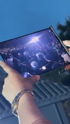 The video shows a crowd of people, mostly middle-aged and older men, standing outside a building at dusk.  Many are holding up their phones, apparently recording something in front of them. The lighting is a bit dim, but there's a bright light illuminating the crowd. Some people are wearing face masks.  The camera moves through the crowd, briefly showing a screen displaying what appears to be a music concert before focusing on the group of people.