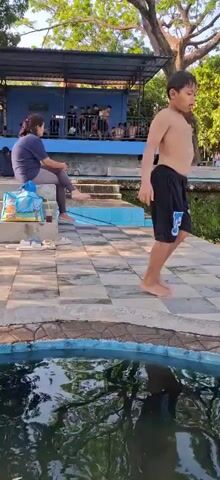 A group of people are gathered around a swimming pool or natural spring. In the foreground, the rippling surface of the water is visible. A person in black shorts and a black t-shirt is walking away from the camera towards the water. Further back, a woman in a dark blue t-shirt and grey leggings sits on a step, with a blue bag and a drink next to her. Beyond her, a set of tiered steps leads up to a shaded area where more people are seated. Statues are visible on higher levels. The environment appears to be outdoors with trees and foliage surrounding the area, suggesting a park or recreational facility. The lighting indicates it is daytime, likely late afternoon given the shadows.