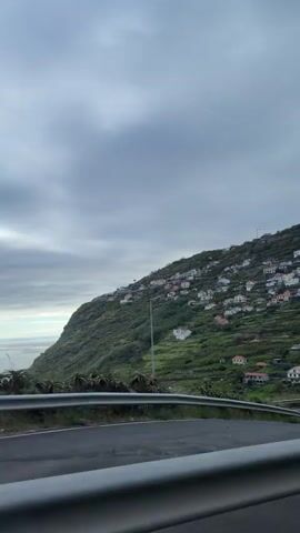 The video shows a lush, green hillside dotted with small, colorful houses. A road runs along the base of the hill, and a gray guardrail is visible in the foreground. The sky is overcast with clouds. The camera appears to be moving along the road, providing a view of the hillside as it passes by. At one point a billboard is seen beside the road.