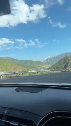 The video shows a view from the front seat of a car driving on a mountain road. The landscape features green, red, and brown hills and mountains, with snow-capped peaks visible in the distance. The sky is blue with scattered clouds. As the car approaches a curve, three figures appear on the side of the road near a guardrail. They appear to be casually standing and looking at the scenery. The road has white dashed lines marking the center and directional signs.