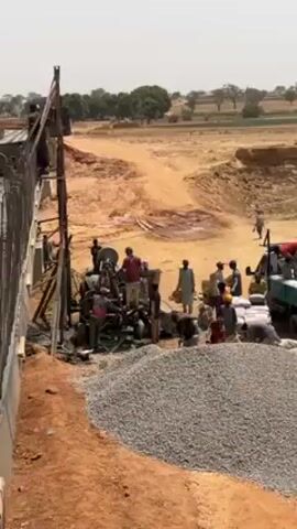 The camera gives a medium shot of a construction site. In the center are many men working to create cement using a cement mixer and other supplies. A large pile of rocks sits directly in front of the camera. To the left, a tall structure is visible, with a man working on it. Behind the men and the cement mixer is a road under construction, and in the background is a rural landscape with some trees. A truck is parked on the right, with bags of cement stacked on the back. The scene is filled with activity as the men work together on the construction project.